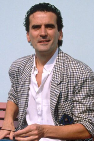massimo troisi nel 1989 fotografato al sole seduto su una panchina sorridente con giacca a quadretti e camicia bianca