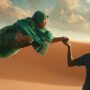 io capitano film fra le dune del deserto una donna africana vestita di verde in volo tiene la mano di un ragazzo senegalese con maglia calcio